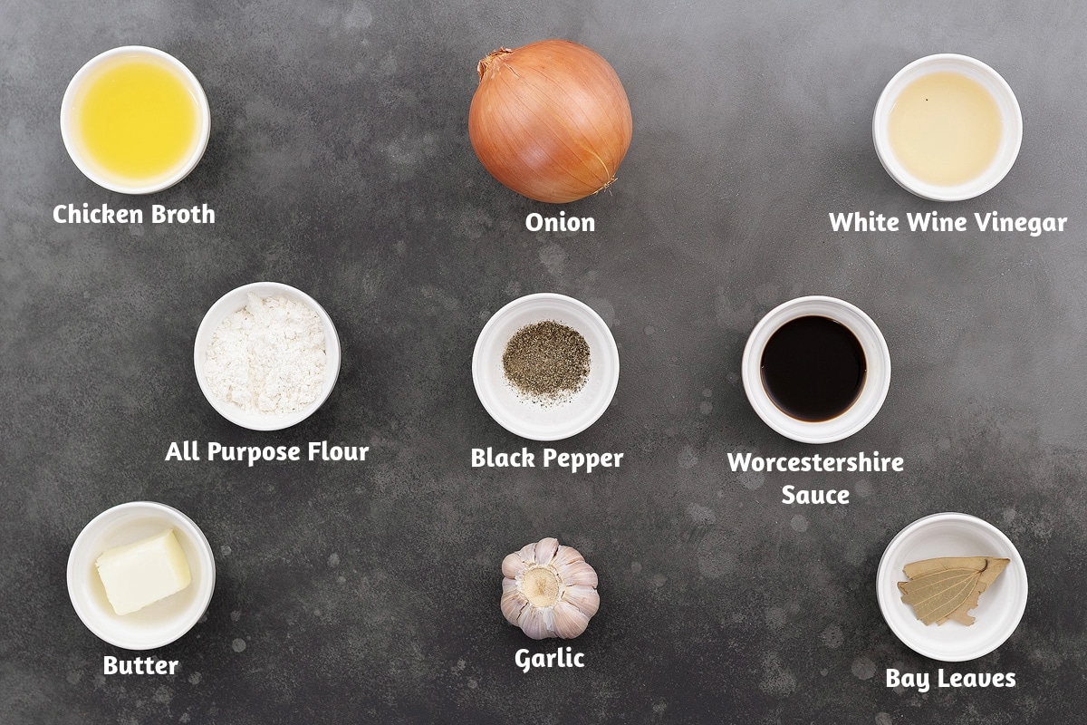 Ingredients for French onion soup arranged on a gray table, including chicken broth, onions, white wine vinegar, all-purpose flour, black pepper powder, Worcestershire sauce, butter, garlic, and bay leaves.