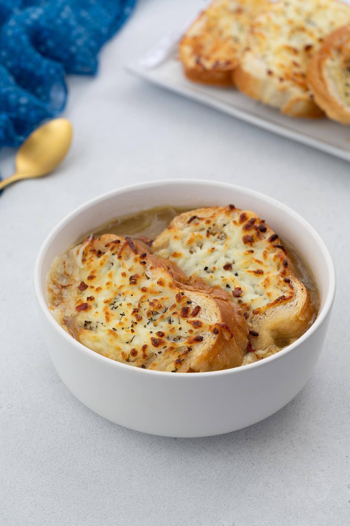 Homemade French onion soup served in a white bowl, garnished with crusty bread on top. The setting includes a golden fork and a blue towel arranged nearby.