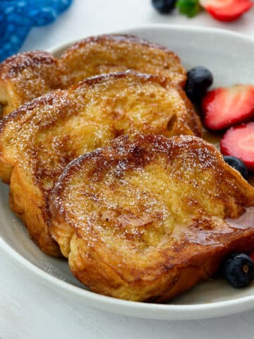 Homemade French toast slices drizzled with syrup and dusted with sugar, served in a white bowl alongside blueberries and strawberries.