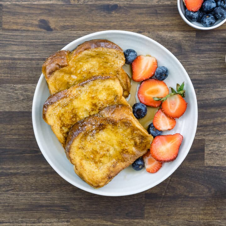French Toast served in a white bowl, topped with syrup, dusted with powdered sugar, and adorned with fresh strawberries and blueberries.