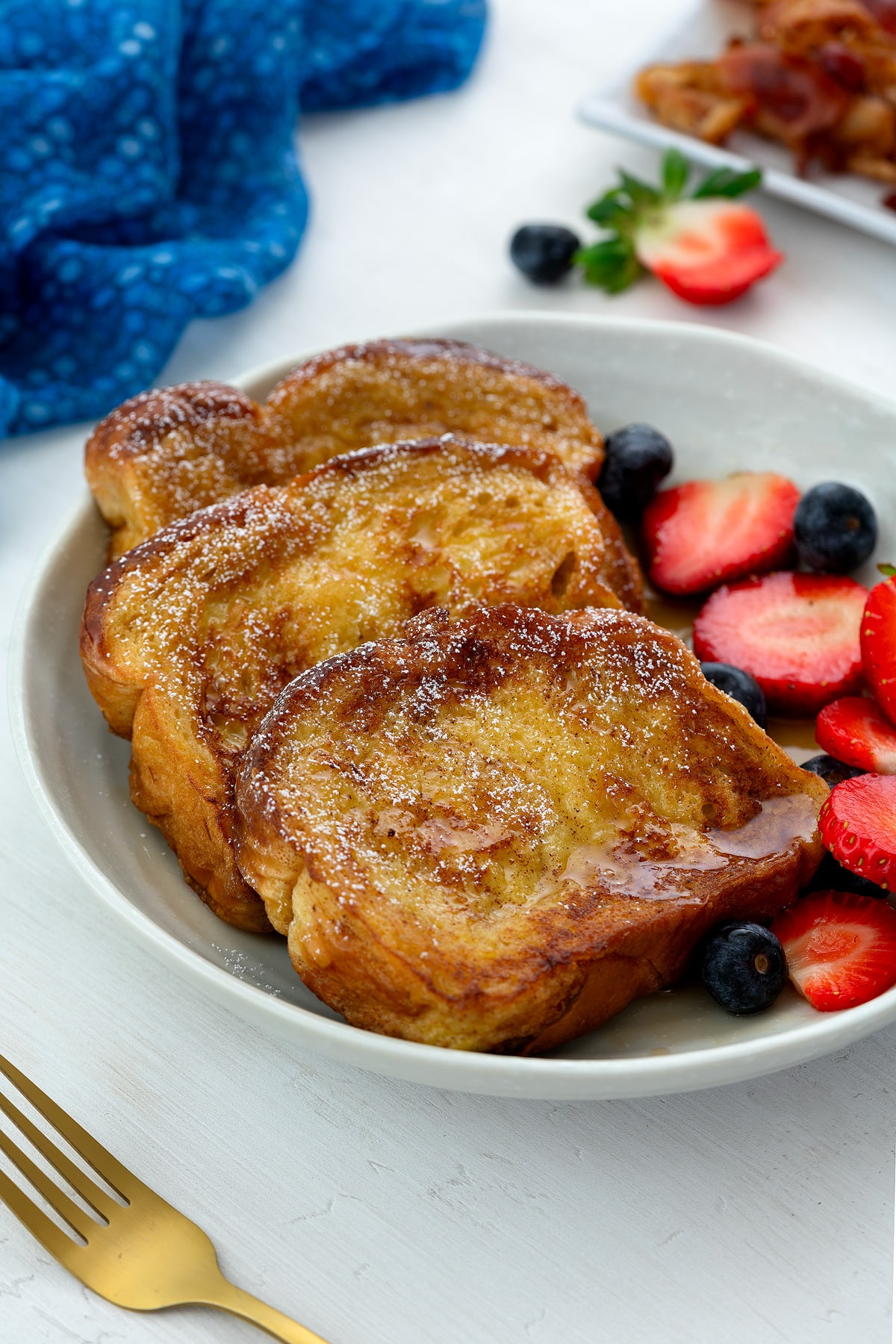 Homemade French toast slices drizzled with syrup and dusted with sugar, served in a white bowl alongside blueberries and strawberries. A blue towel, golden fork, and bacon on a plate are arranged around the bowl.