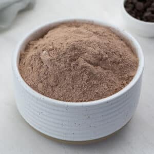 Hot Cocoa Mix in a white bowl on a white table, surrounded by a cup of chocolate chips and a towel.