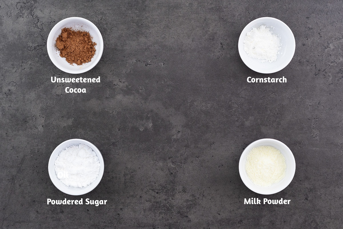 Hot Cocoa Mix Recipe Ingredients arranged on a gray table: Unsweetened Cocoa, Cornstarch, Powdered Sugar, Milk Powder.