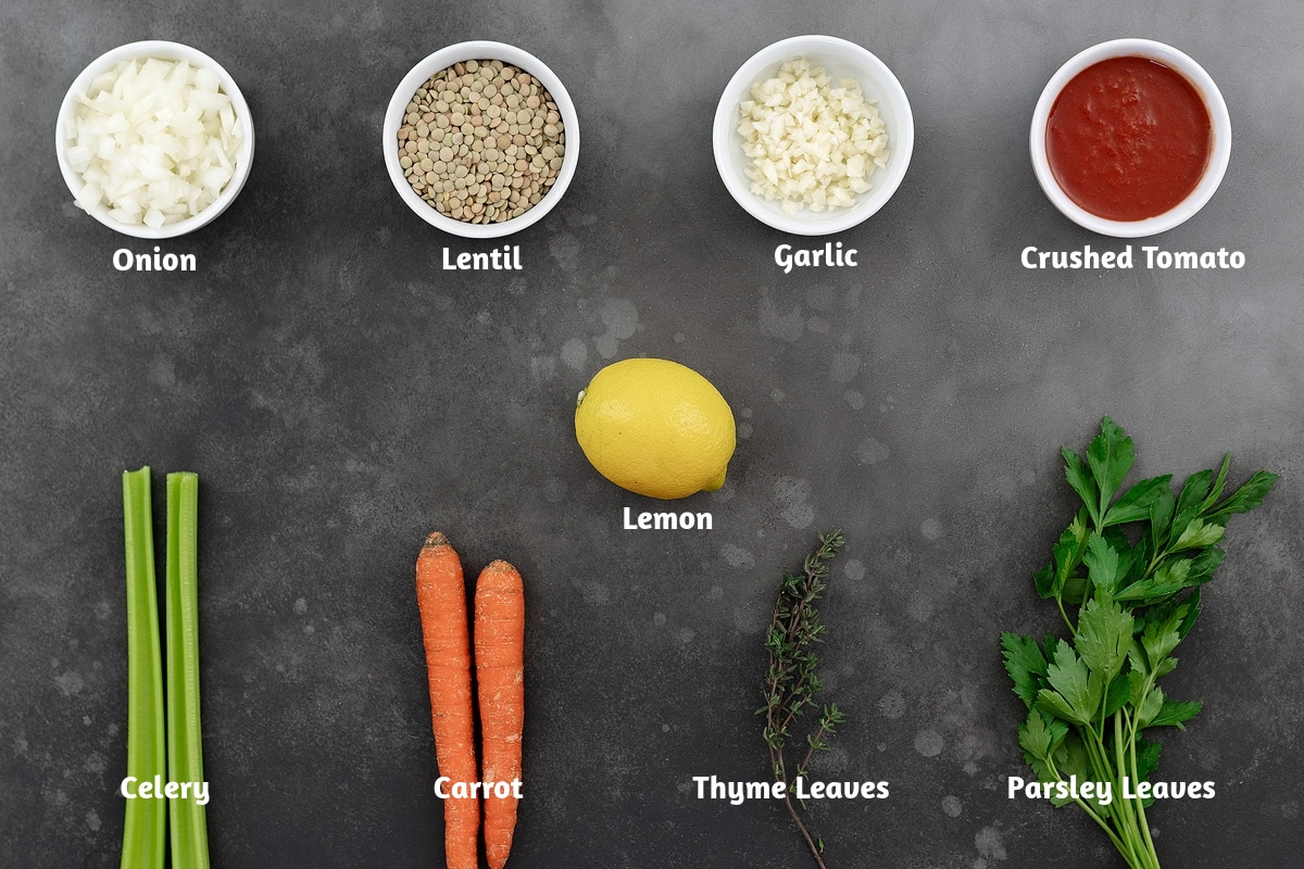 Ingredients for lentil soup on a gray table: onion, lentils, garlic, tomatoes, celery, carrot, lemon, thyme, and parsley.