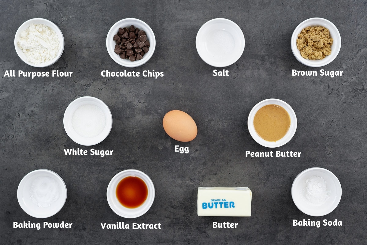 Ingredients for peanut butter chocolate chip cookies - flour, chocolate chips, salts, sugars, egg, peanut butter, baking powder, vanilla, butter, baking soda - arrayed on a gray table.