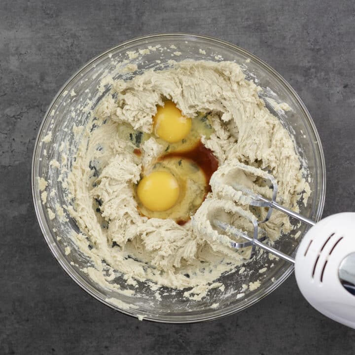 Creamy butter-sugar mixture in a bowl with eggs and vanilla extract, being blended with an electric beater.