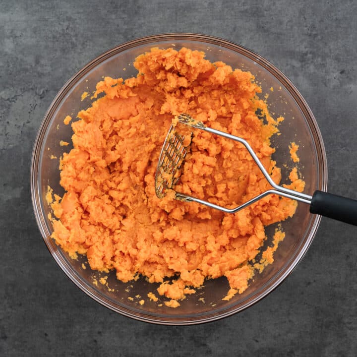 Bowl with mashed sweet potatoes, accompanied by a potato masher on the side.