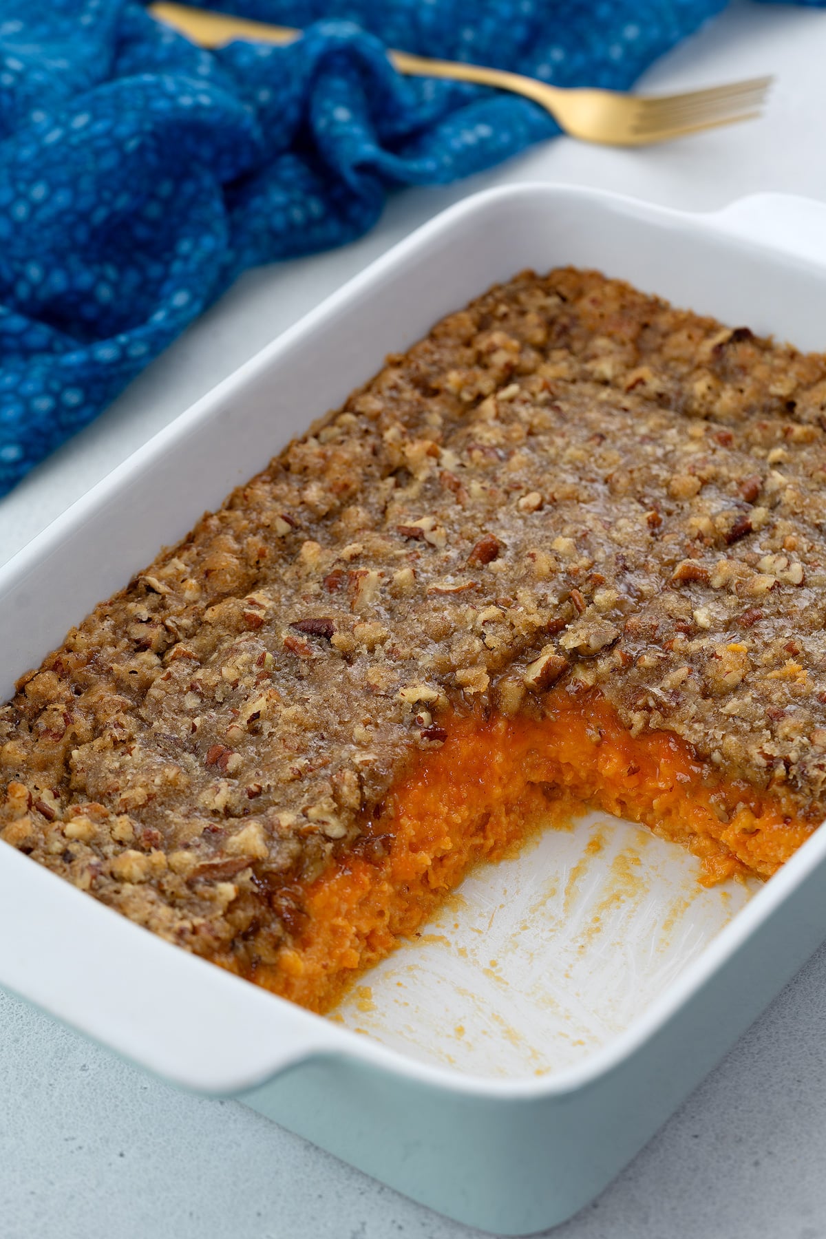 Sweet potato casserole in a white baking dish on a table, with a slice taken out, accompanied by a blue towel and a golden fork.