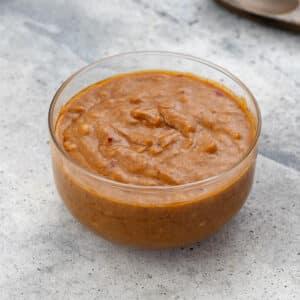 Thai peanut sauce in a glass bowl on a white surface, with a wooden spoon nearby.