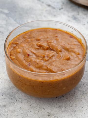 Thai peanut sauce in a glass bowl on a white surface, with a wooden spoon nearby.