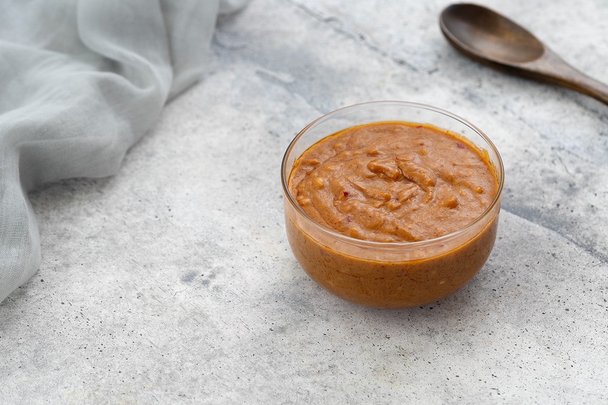 Thai peanut sauce in a glass bowl on a white surface, with a towel and a wooden spoon nearby.
