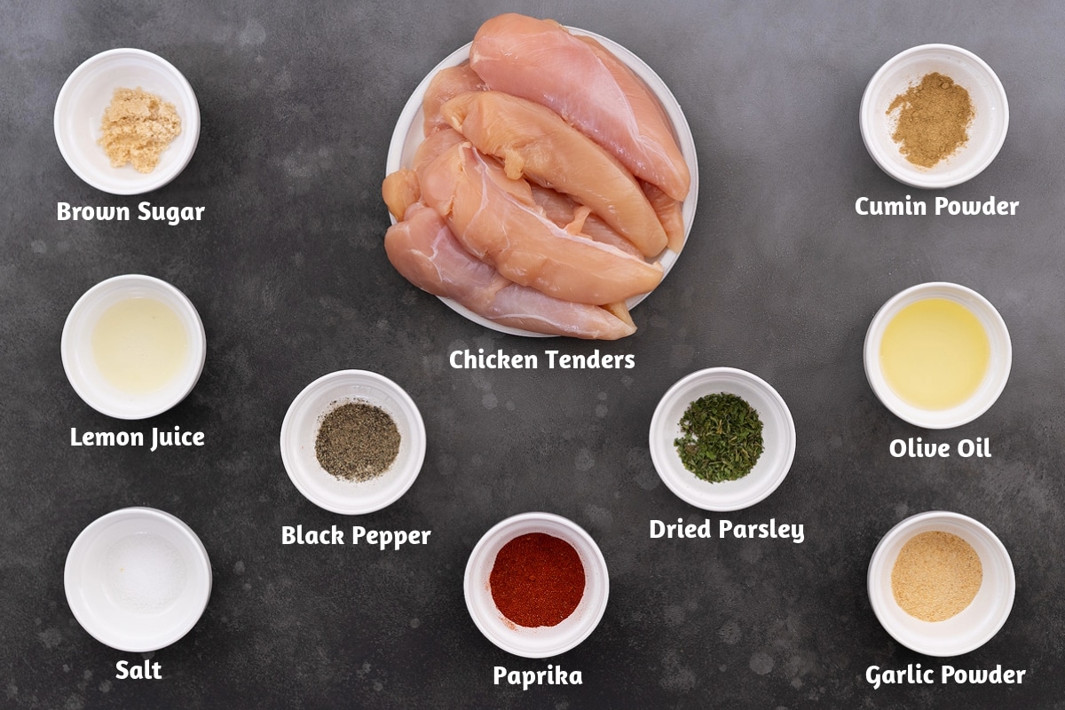 Ingredients for baked chicken tenders recipe on a grey table, including brown sugar, cumin powder, lemon juice, black pepper, dried parsley, olive oil, salt, paprika, and garlic powder.