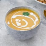 Homemade butternut squash soup in a white bowl on a white table, with a bowl of farro, and a golden spoon nearby.