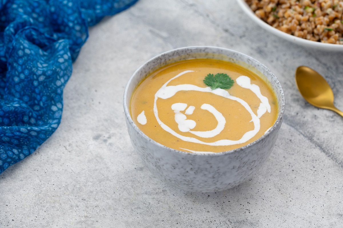 Homemade butternut squash soup in a white bowl on a white table, with a blue towel, a bowl of farro, and a golden spoon nearby.