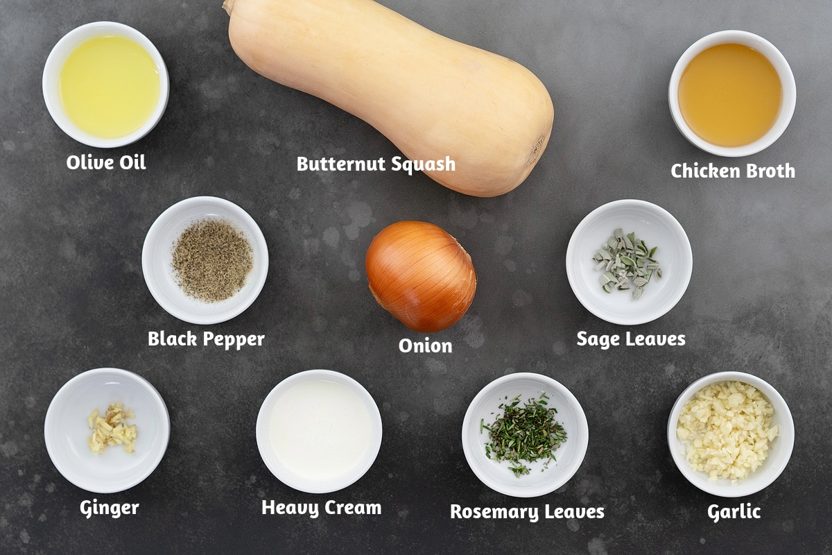Butternut squash soup recipe ingredients laid out on a gray table, including olive oil, butternut squash, chicken broth, black pepper powder, onion, sage leaves, ginger, heavy cream, rosemary leaves, and garlic.