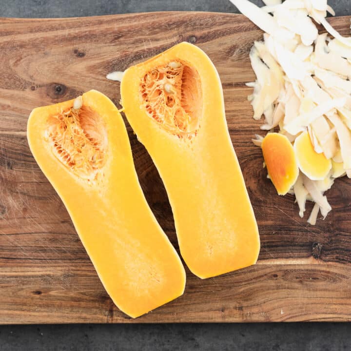 A wooden board with vertically halved butternut squash and its peels alongside.