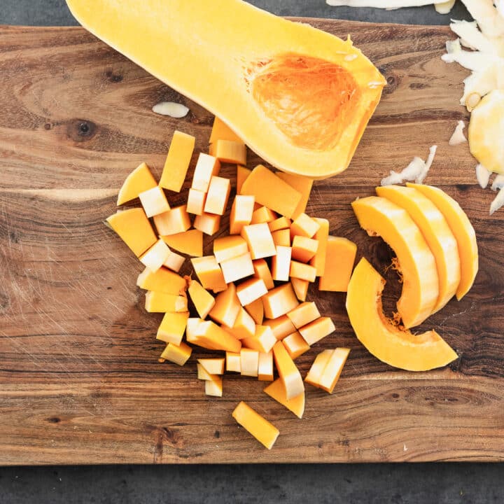 A wooden board with bite-sized chopped butternut squash, with the other unsliced half on the side.
