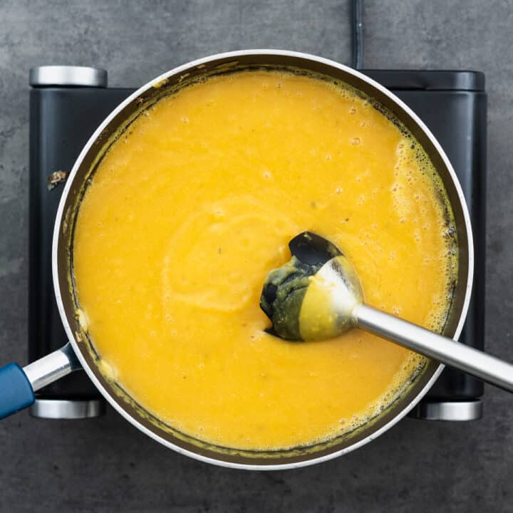A pan with blended creamy butternut squash soup, with an immersion blender on the side.