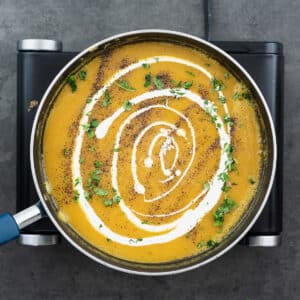 A pan with velvety butternut squash soup garnished with cream, black pepper, and cilantro leaves.