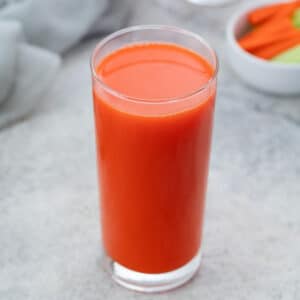 A glass of fresh carrot juice on a white table, next to a bowl of chopped carrots and celery, with a towel placed around.