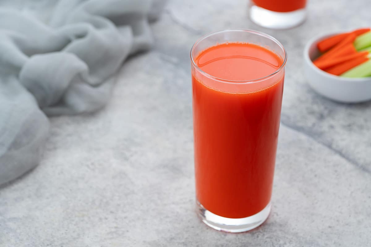 A glass of fresh carrot juice on a white table, next to a bowl of chopped carrots and celery, with a towel placed around.