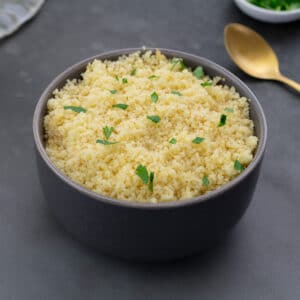 Cooked couscous in a grey bowl on a grey table, with a gold fork, parsley in a small cup, and a towel arranged around it.