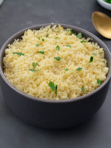 Cooked couscous in a grey bowl on a grey table, with a gold fork, parsley in a small cup, and a towel arranged around it.