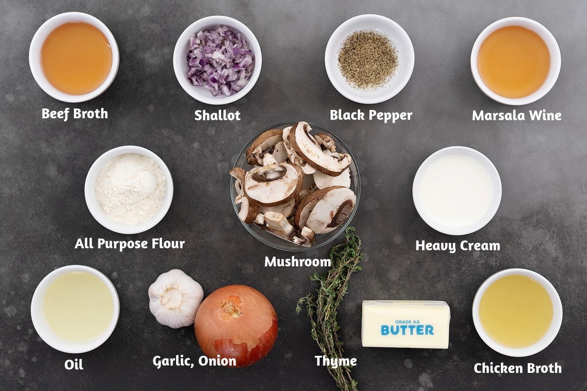 Ingredients for cream of mushroom soup arranged on a grey table, including beef broth, shallots, black pepper, Marsala wine, all-purpose flour, mushrooms, heavy cream, oil, garlic, onion, thyme, butter, and chicken broth.