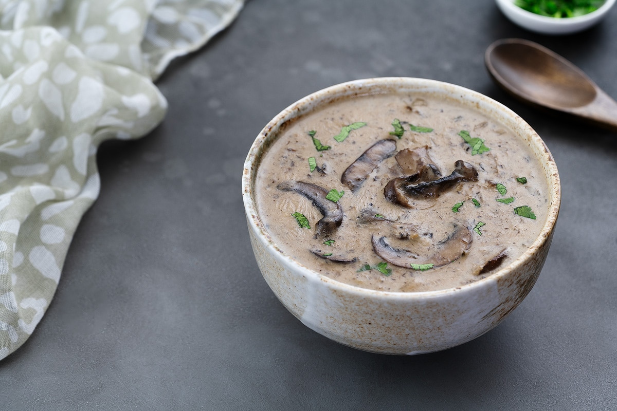 Cream of mushroom soup in a cup on a gray table, with a wooden spoon, towel, and cilantro in a small cup nearby.