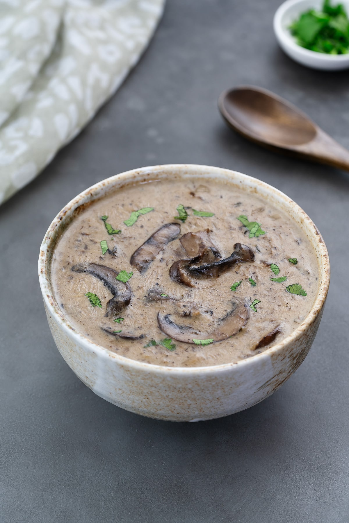 Cream of mushroom soup in a cup on a gray table, with a wooden spoon, towel, and cilantro in a small cup nearby.