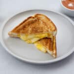 Grilled cheese sandwich in a white bowl on a table, with a cup of tomato soup nearby.