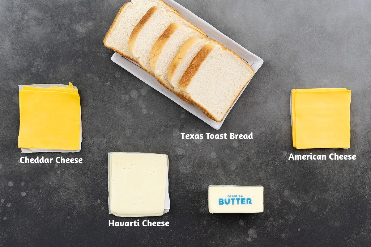Ingredients for a Grilled Cheese Sandwich, including Texas Toast Bread, Cheddar and American Cheese slices, Havarti Cheese, and Butter, arranged on a gray table.
