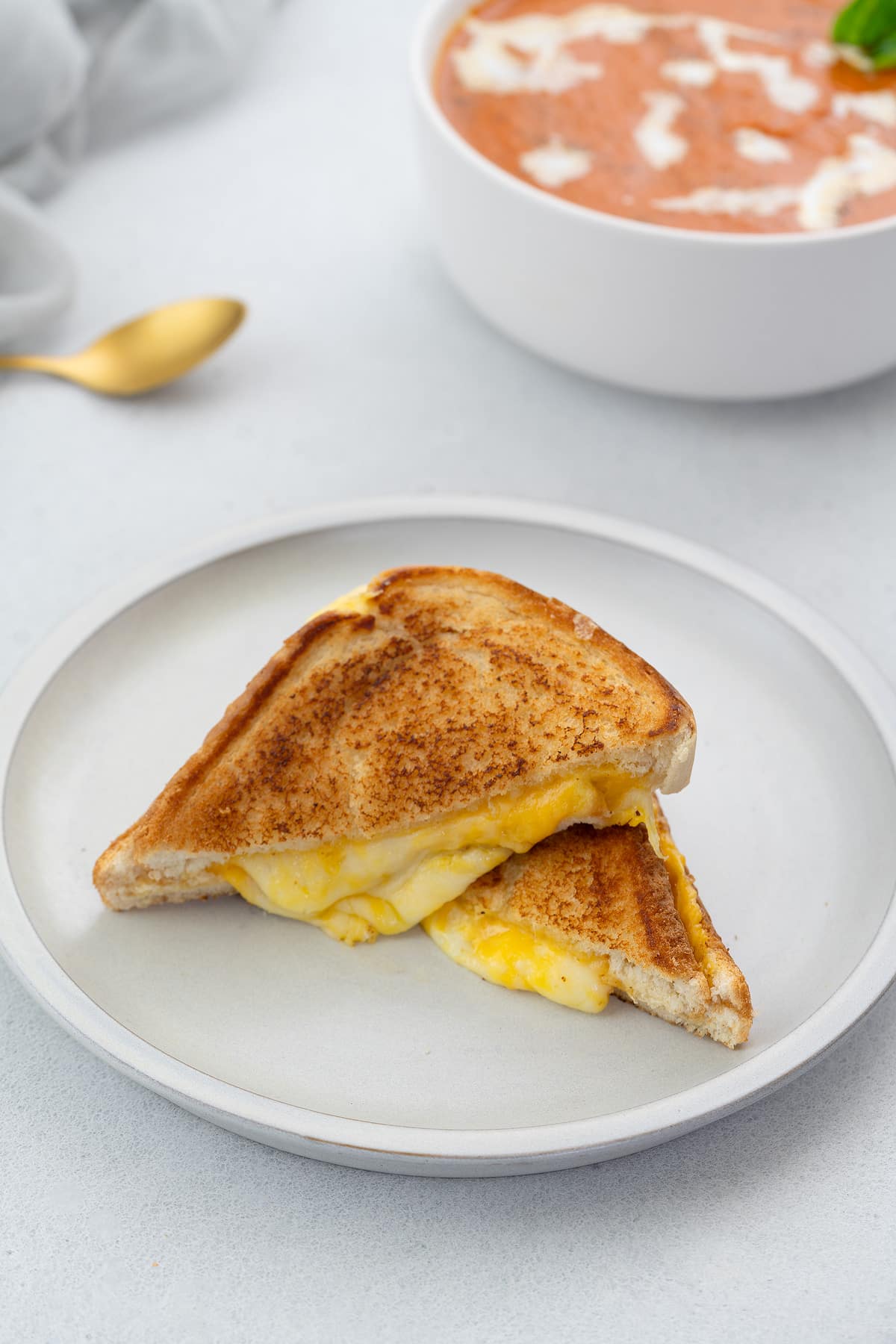 Grilled cheese sandwich in a white bowl on a table, with a golden spoon and a cup of tomato soup nearby.