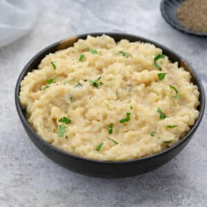 Creamy risotto served in a grey bowl on a white table, accompanied by pepper powder in a small container, and a white towel arranged nearby.