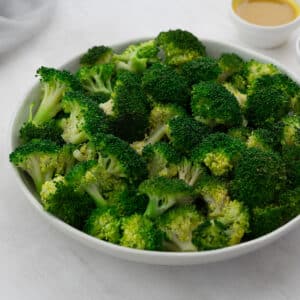 Steamed broccoli in a white bowl on a white table, with honey mustard sauce in nearby small bowl.