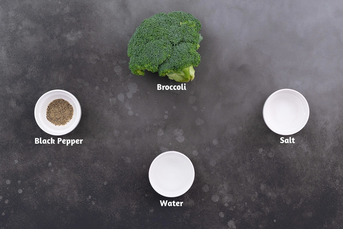 Fresh broccoli, black pepper, and salt arranged on a grey table, with a cup of water nearby.