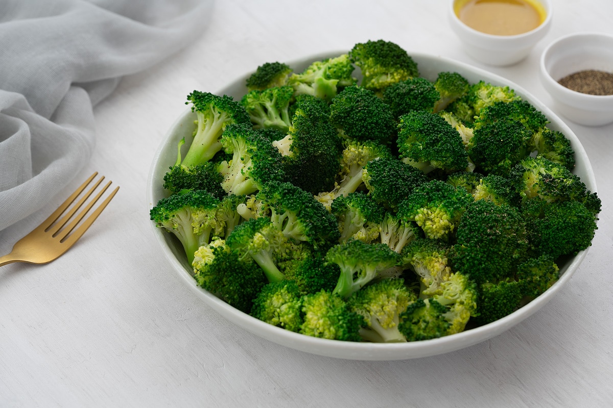 Steamed broccoli in a white bowl on a white table, with honey mustard sauce and pepper in nearby small bowls.