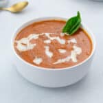 Creamy tomato soup served in a white bowl on a white table, accompanied by a golden fork nearby.
