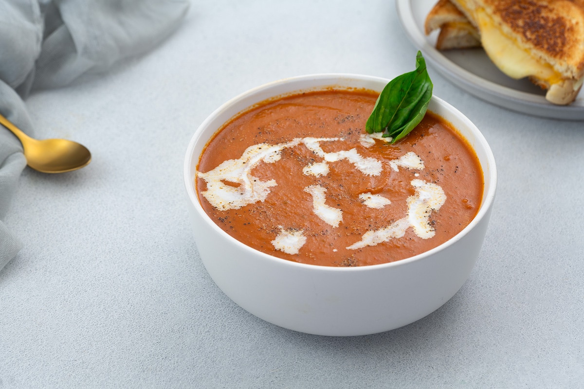 Creamy tomato soup served in a white bowl on a white table, accompanied by grilled cheese and a golden fork, with a towel nearby.