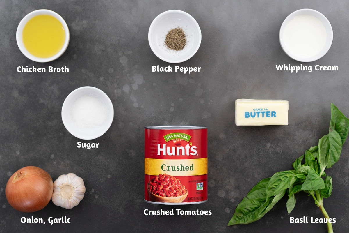 Ingredients for tomato soup displayed on a gray table, including chicken broth, black pepper, whipping cream, sugar, butter, onion, garlic, crushed tomatoes, and basil leaves.