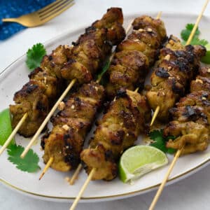 Homemade chicken satay on skewers, served on a plate garnished with lime wedges and cilantro leaves. Beside the plate, there is a golden fork, and a blue towel.