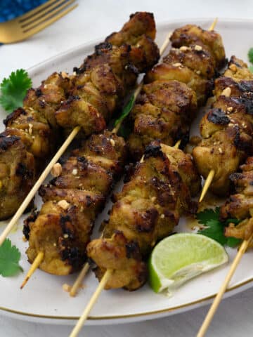 Homemade chicken satay on skewers, served on a plate garnished with lime wedges and cilantro leaves. Beside the plate, there is a golden fork, and a blue towel.