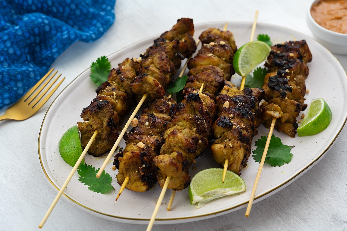 Homemade chicken satay on skewers, served on a plate garnished with lime wedges and cilantro leaves. Beside the plate, there is a golden fork, a blue towel, and a cup of peanut sauce.