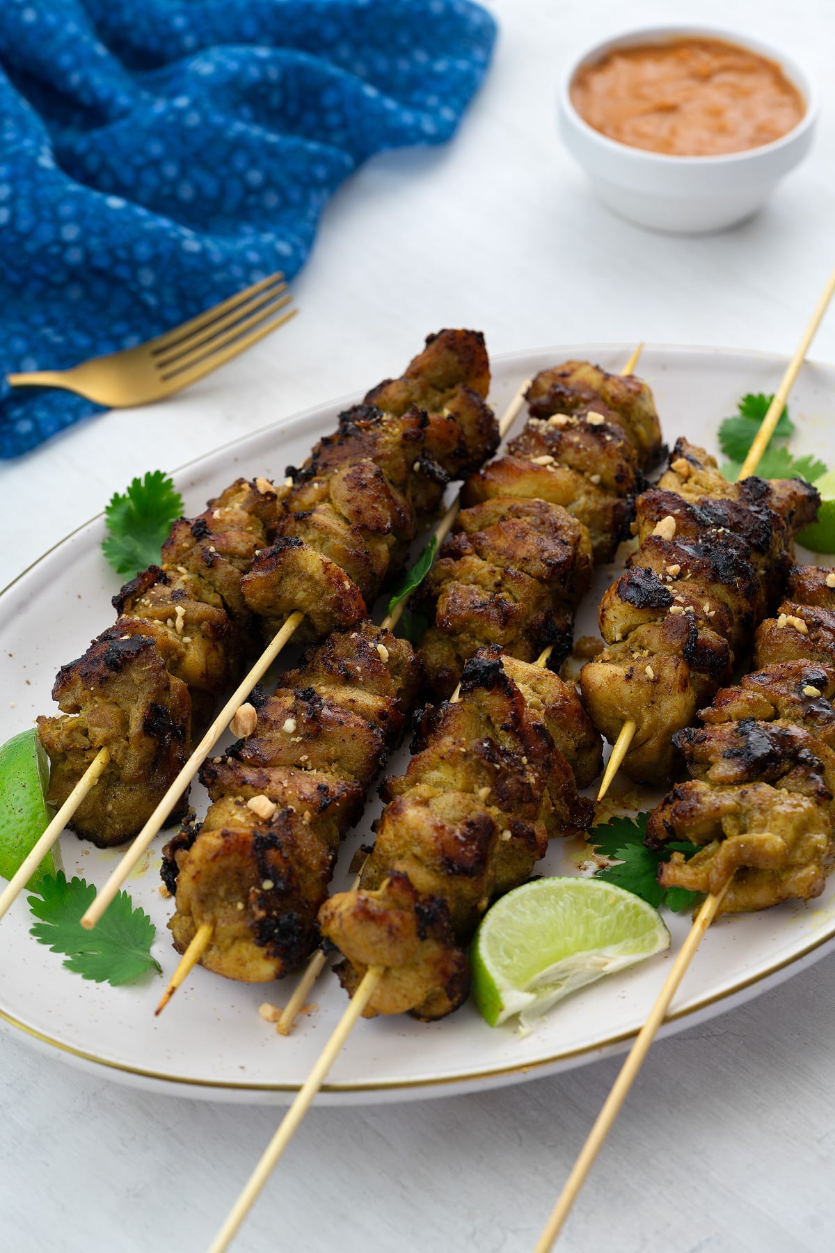 Homemade chicken satay on skewers, served on a plate garnished with lime wedges and cilantro leaves. Beside the plate, there is a golden fork, a blue towel, and a cup of peanut sauce.