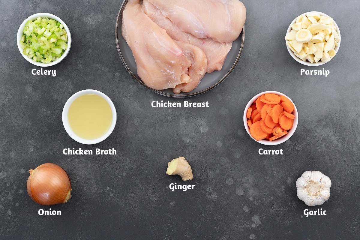 Ingredients for chicken soup arranged on a grey table: celery, chicken breast, and parsnip in the first group; chicken broth, ginger, and carrot in the second; onion and garlic in the third.