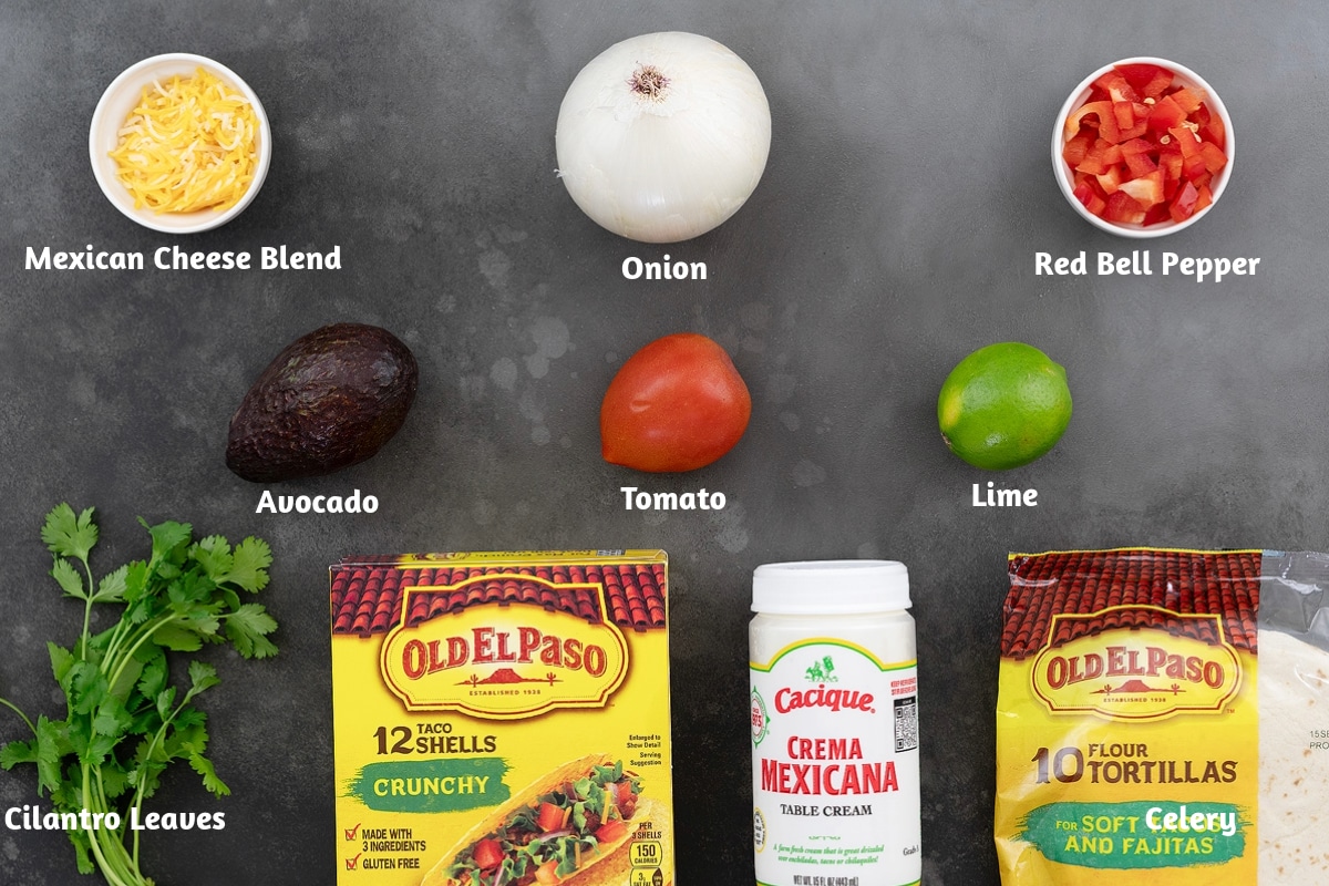 On a gray table, the ingredients for chicken tacos are neatly arranged, including Mexican cheese blend, onion, red bell pepper, avocado, tomato, lime, cilantro leaves, taco shells, table cream, and tortillas.