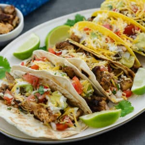 Chicken Taco served in a white plate with lime wedges and cilantro leaves.