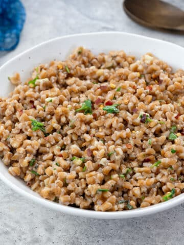 Cooked farro in a white bowl on a white table, with a wooden spoon, and a blue towel nearby.