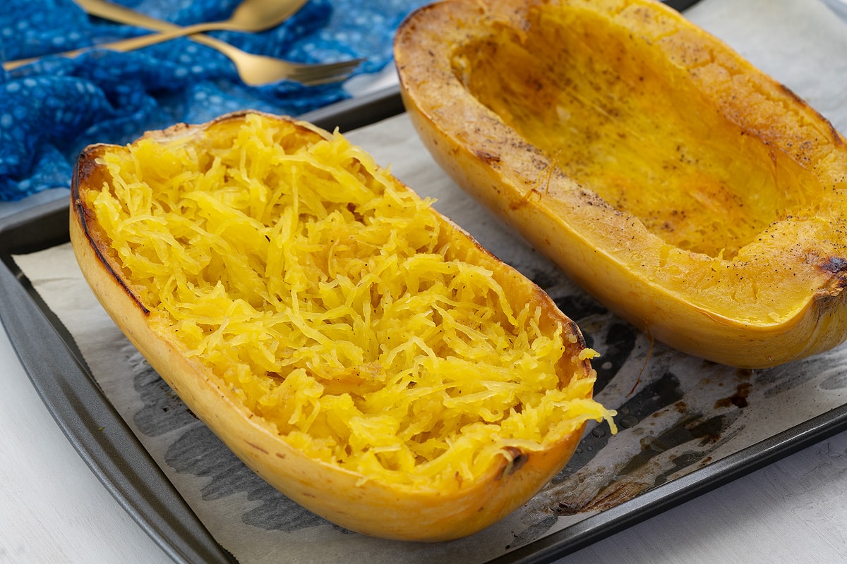 Two halves of baked spaghetti squash on a tray, with a blue towel and a golden fork nearby.