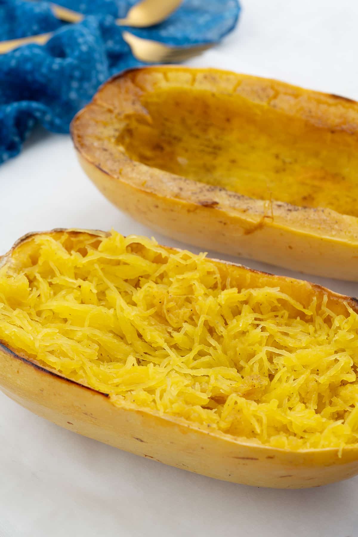 Two halves of baked spaghetti squash on a white table, with a blue towel and a golden fork nearby.